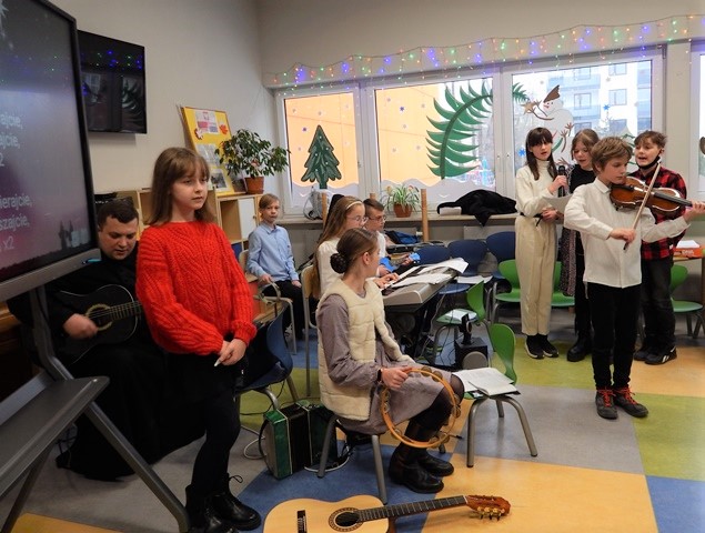 Uczniowie z klasy 7b i Ksiądz Krzysztof Wiśniewski grający na instrumentach muzycznych i śpiewający kolędy, tablica multimedialna z tekstami kolęd