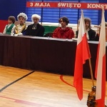 Uczniowie siedzący za stołem, ubrani tak jak ludzie z XVIII wieku, za nimi napis - 3 Maja Święto Konstytucji. Po prawej stronie dwie flagi biało czerwone