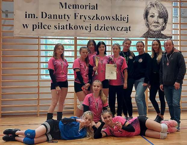 Dziewczęta z drużyny siatkarskiej z naszej szkoły razem z trenerem panem Krzysztofem Kubikiem na tle baneru z wizerunkiem pani Danuty Fryszkowskiej