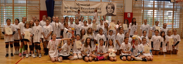 Dziewczynki wraz z trenerami biorące udział w Memoriale, nad nimi baner z nazwą Memoriału