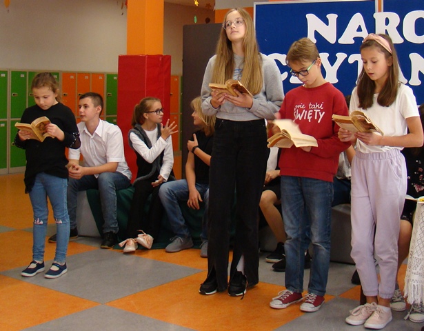 Uczniowie czytający utwór, za nimi tablica z napisem Narodowe Czytanie w 33. Na niej życiorys Adama Mickiewicza